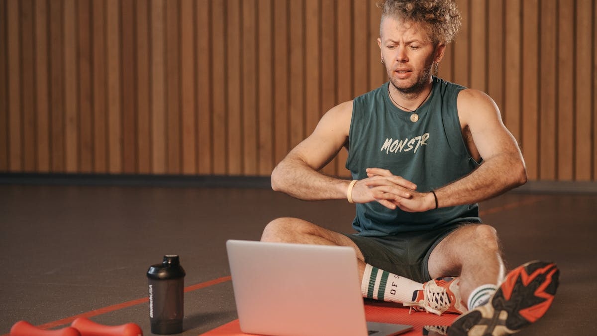 Adult man exercising indoors with a laptop for virtual fitness training, sitting on a red mat.